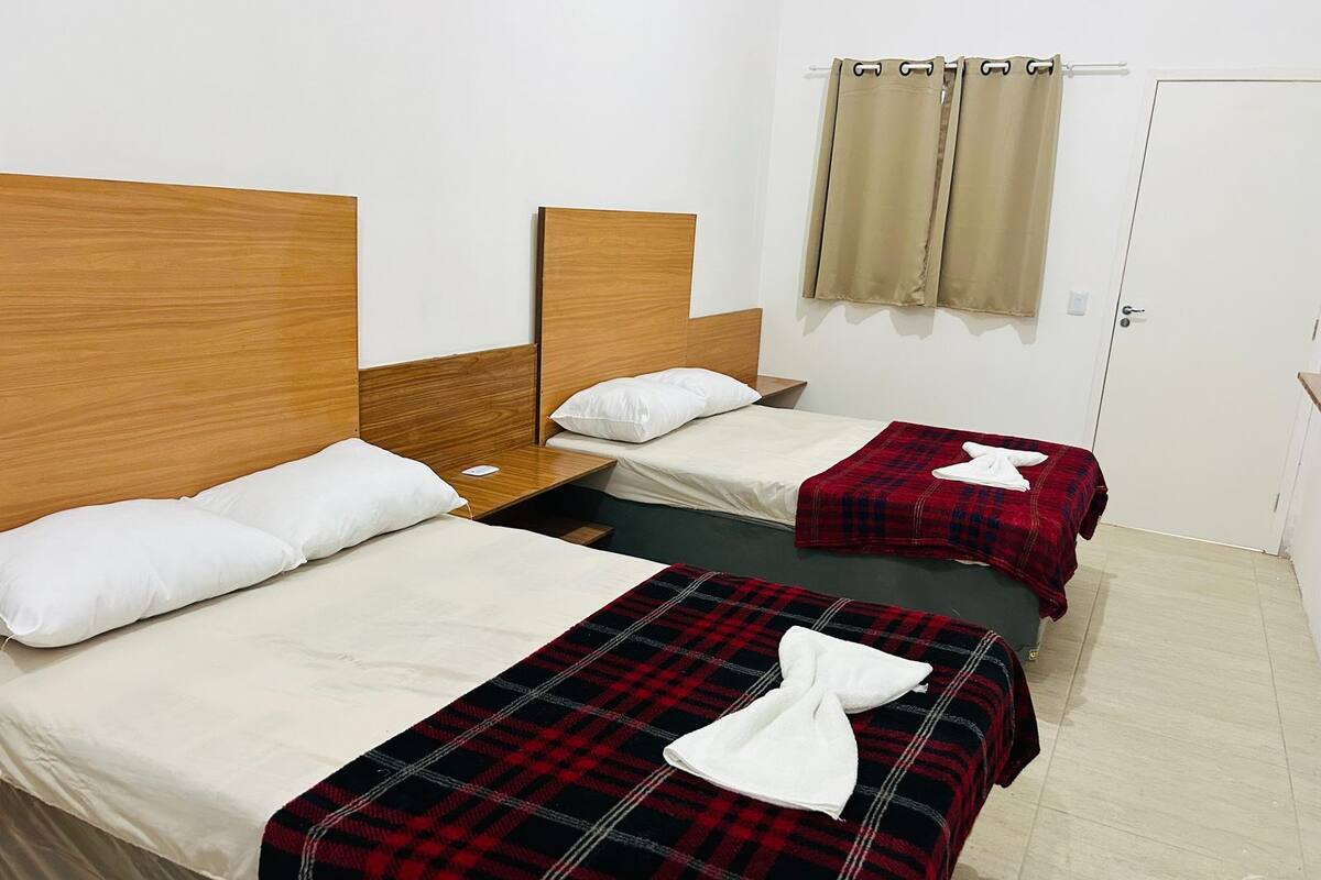 Habitación de hotel con dos camas, cada una con cabecero de madera, ropa de cama blanca y manta de cuadros rojos. Se observa una mesita de noche con lámpara y toma de corriente.