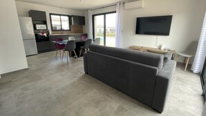 Una sala de estar moderna con un sofá gris, una zona de comedor con mesa y sillas de vidrio, una cocina con gabinetes grises y un televisor montado en la pared.