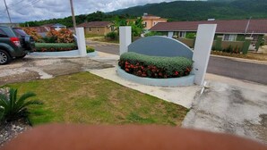 A residential area with a paved driveway, a green SUV parked on the left, and a landscaped garden with red flowers in the center.