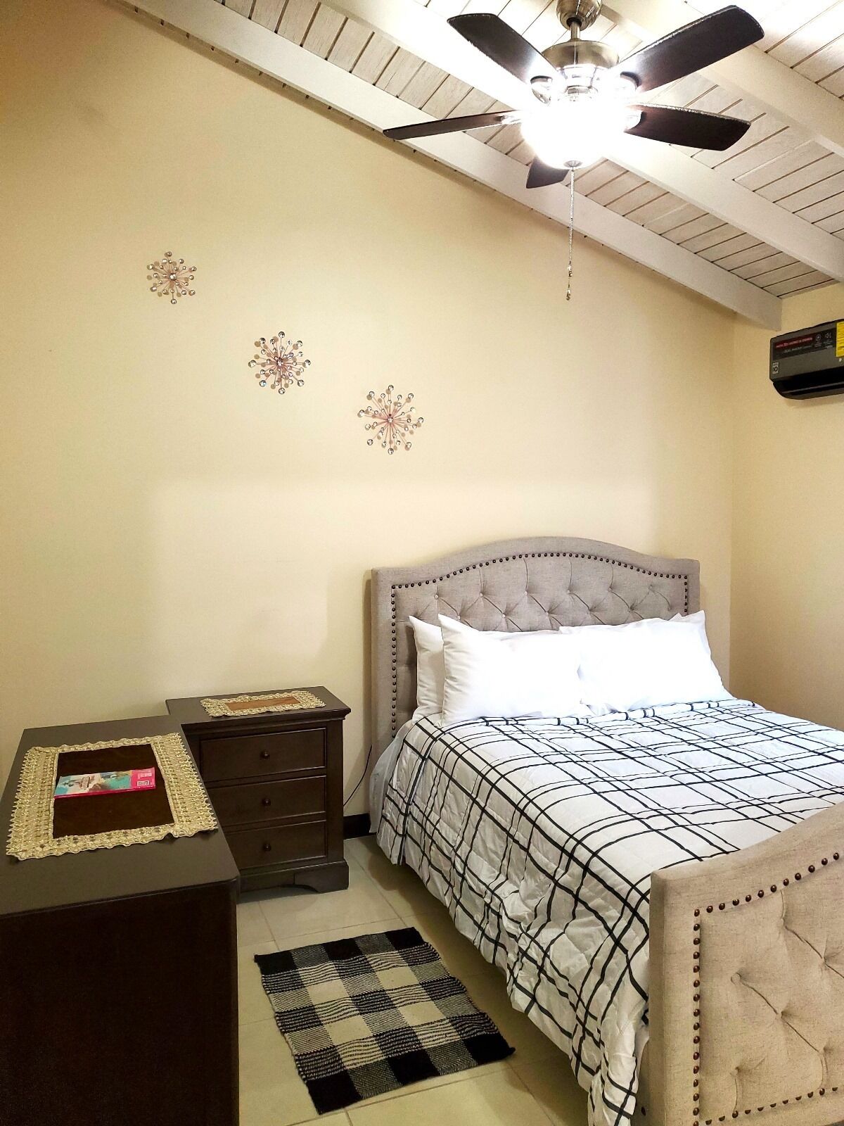 Een slaapkamer met een gestoffeerd hoofdeinde, een bed met een geruit sprei, een nachtkastje met een decoratief object, een donkere dressoir, een plafondventilator en wanddecoraties.