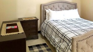 A bedroom with a tufted headboard, a bed with a checkered bedspread, a bedside table with a decorative item, a dark dresser, a ceiling fan, and wall decorations.