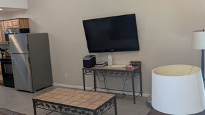 A living room with a brown leather couch, a coffee table, a TV, a lamp, and a kitchen area with a refrigerator and oven. - Barndo in the Rocky Mt Woods (Rocky Mount)