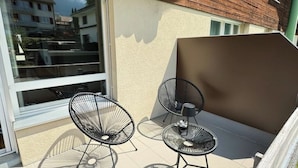 Un balcon avec deux chaises en métal et une petite table, une porte-fenêtre coulissante et une vue sur les immeubles voisins.