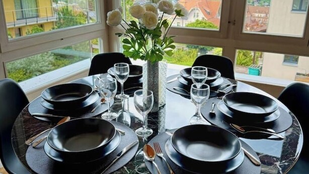 Une table à manger dressée pour six personnes, avec des assiettes noires, des couverts en argent et des verres à vin, entourée de chaises.