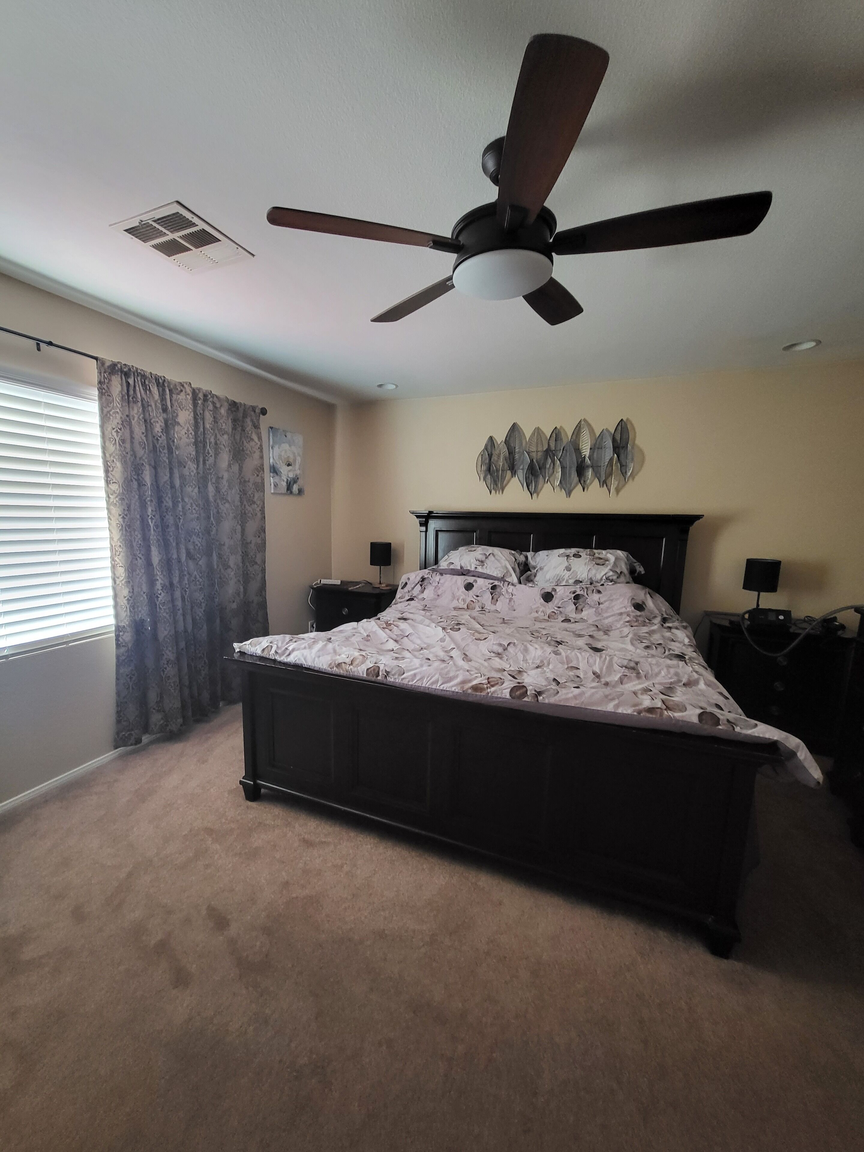 A bedroom with a bed, ceiling fan, wall art, and a window with curtains.
