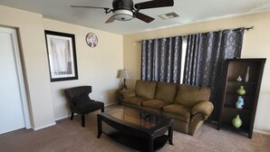 Un salon avec un ventilateur de plafond, un canapé marron, une table basse en verre, un fauteuil noir, une lampe et une étagère décorative.