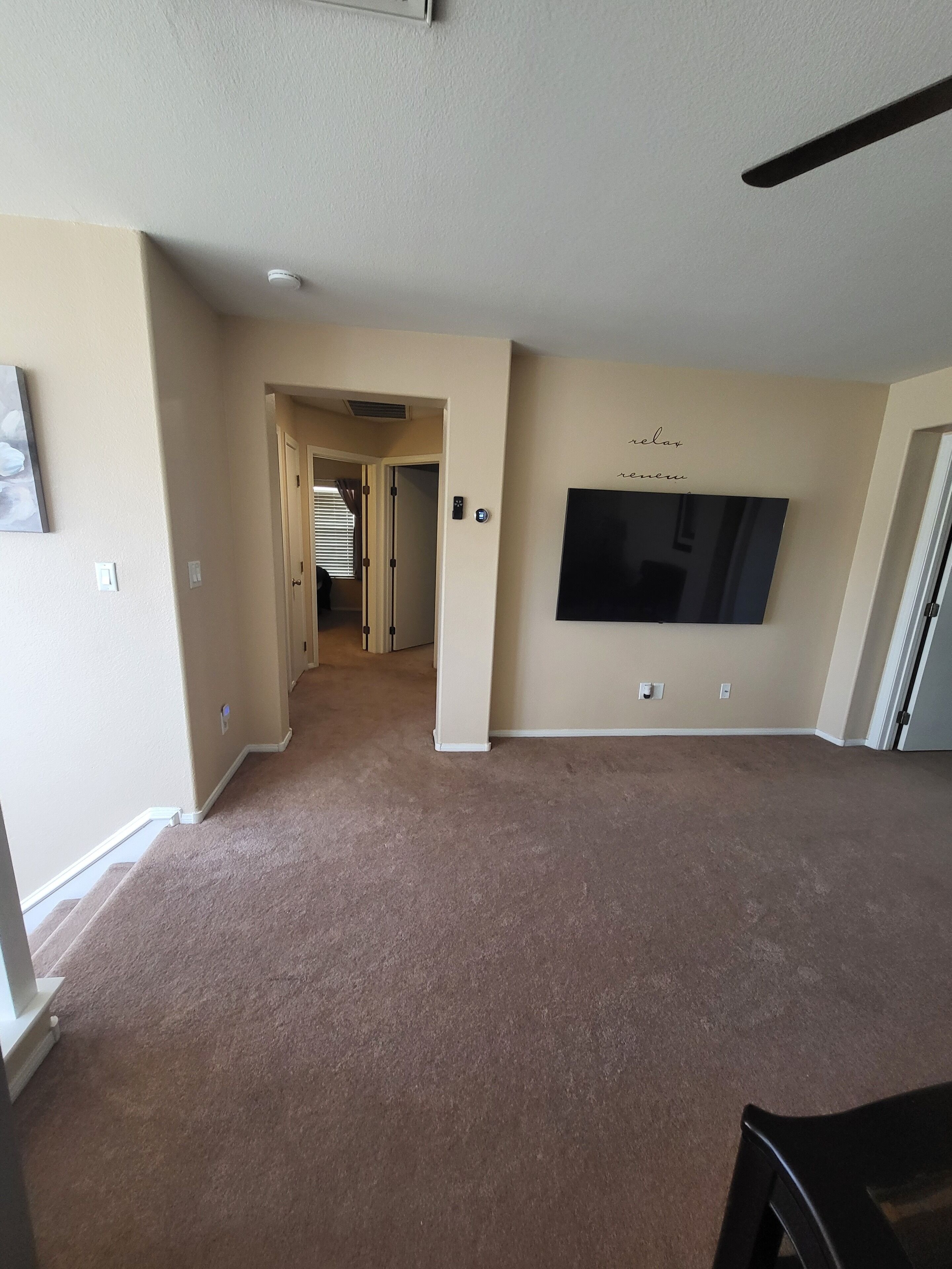 A spacious living area with a flat-screen TV mounted on the wall, carpeted flooring, and a ceiling fan.