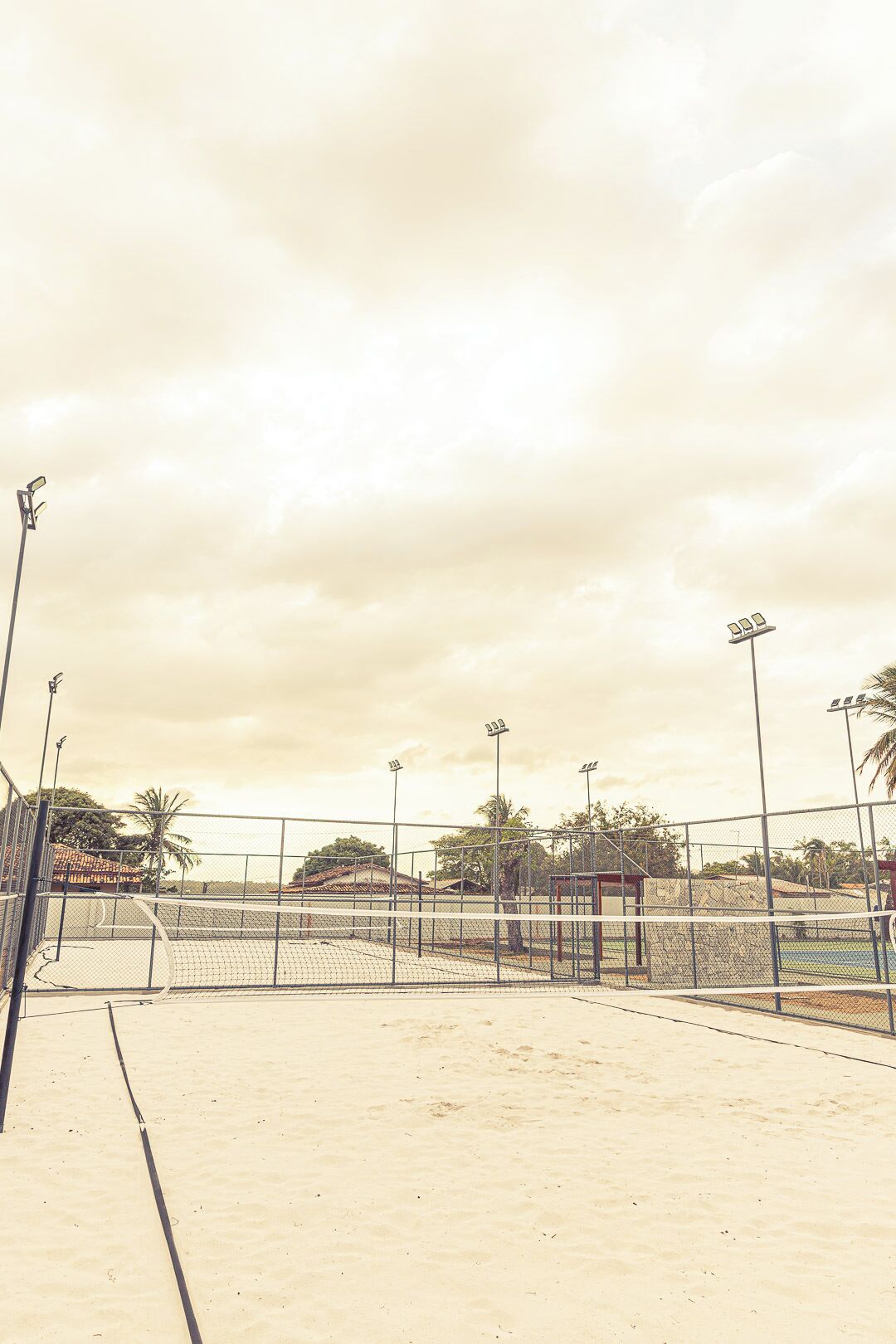 Een buiten tennisbaan met net, omheind door een hek en voorzien van verlichting.