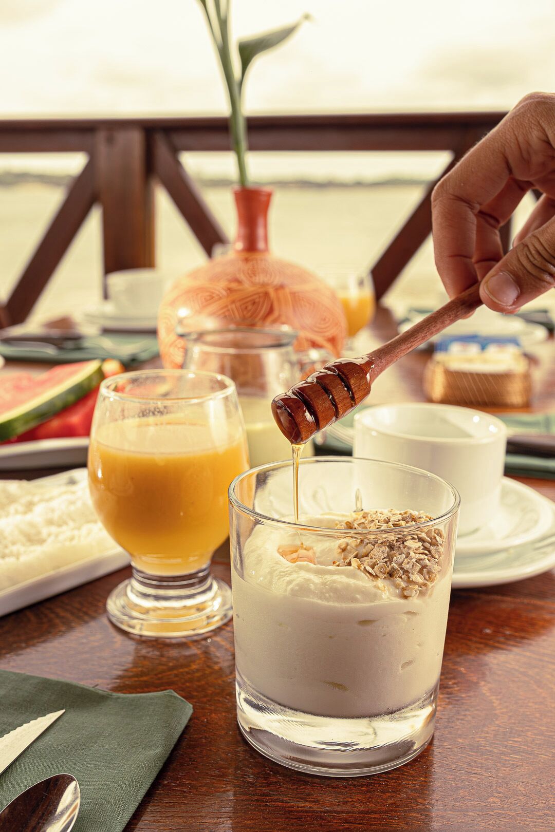 A breakfast setting with a glass of orange juice, a glass of yogurt topped with granola and honey, and a honey dipper in use.