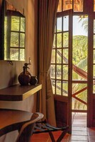 Una habitación con un escritorio de madera, una silla, un ventanal con cortinas y vistas a palmeras al exterior.