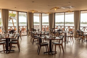 Un restaurante con varias mesas y sillas, ventanas amplias con cortinas y lámparas colgantes.