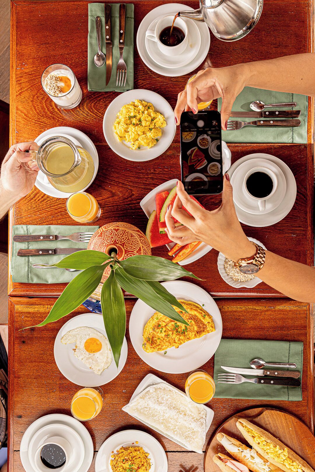 Une table de salle à manger en bois, sur laquelle sont disposés divers aliments pour le petit-déjeuner, notamment des œufs, des toasts et du jus, et une personne prenant une photo avec un smartphone.