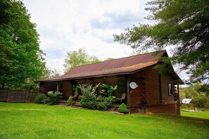 A rustic log cabin with a porch, surrounded by lush greenery and trees.