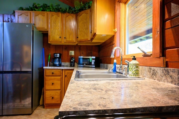 A kitchen with wooden cabinets, a stainless steel refrigerator, a granite countertop, a sink, a faucet, a microwave, and a coffee maker.