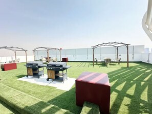 A rooftop terrace with artificial grass, two barbecue grills, outdoor seating, and pergolas providing shade.