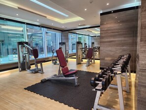 Un gimnasio con equipamiento de ejercicio, incluyendo bancos y mancuernas, y ventanas amplias.