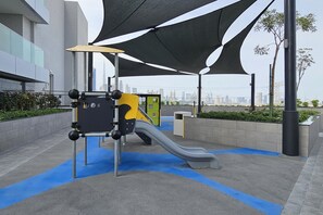An outdoor playground with a slide and climbing structure, surrounded by modern buildings and a view of the city skyline.