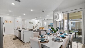 Une cuisine moderne avec des meubles blancs, un îlot central et un coin repas agrémenté d’une table blanche dressée pour six personnes.