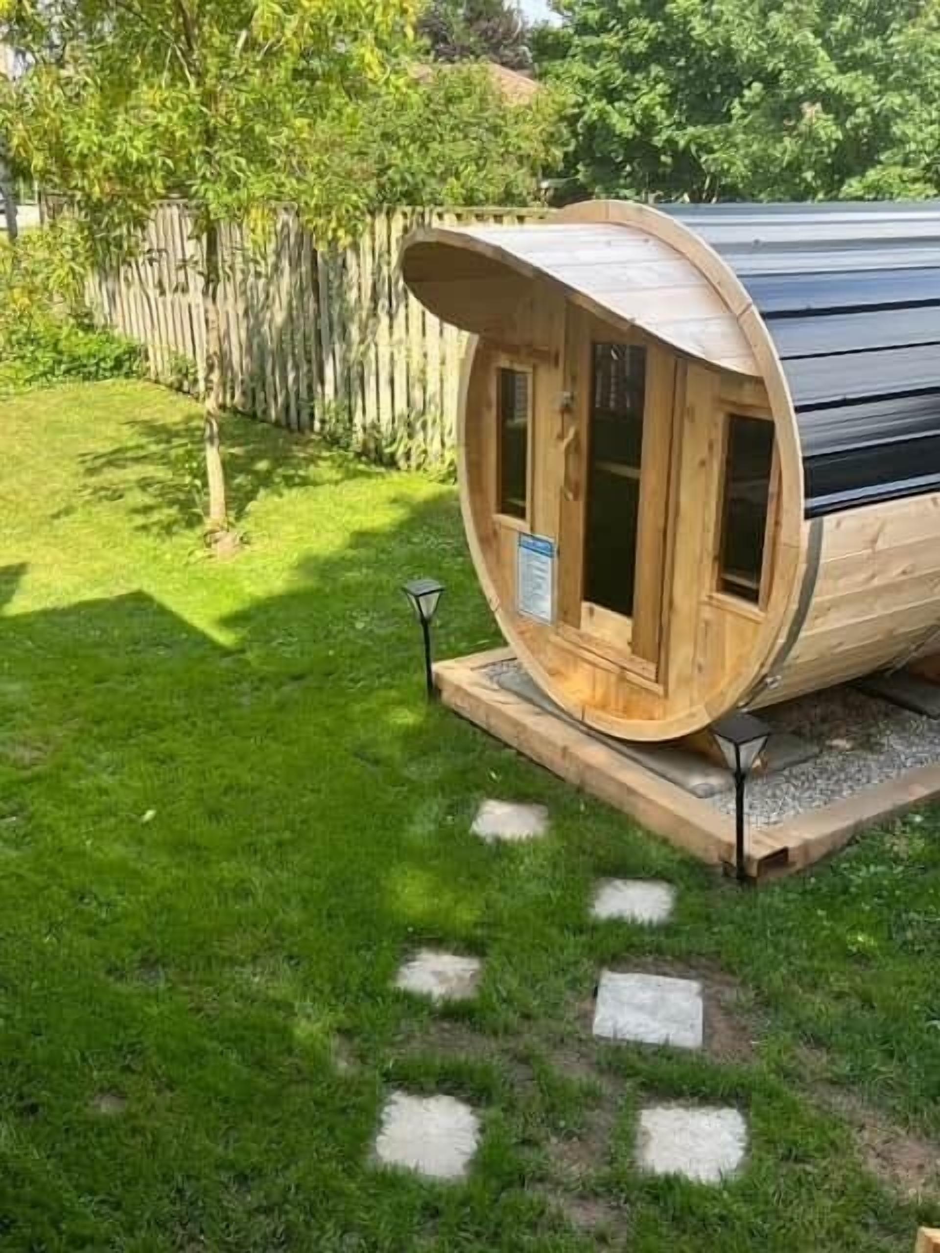 A wooden cylindrical structure with a flat roof, possibly a sauna or hot tub, on a grassy area with a wooden fence in the background.