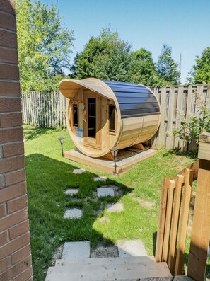 A wooden cylindrical structure with a door and windows, situated in a backyard with a brick wall, wooden fence, and grass. - Walkerton Sauna Suite (Walkerton)