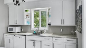 A modern kitchen with white cabinets, a microwave, a sink, and a window with greenery outside.