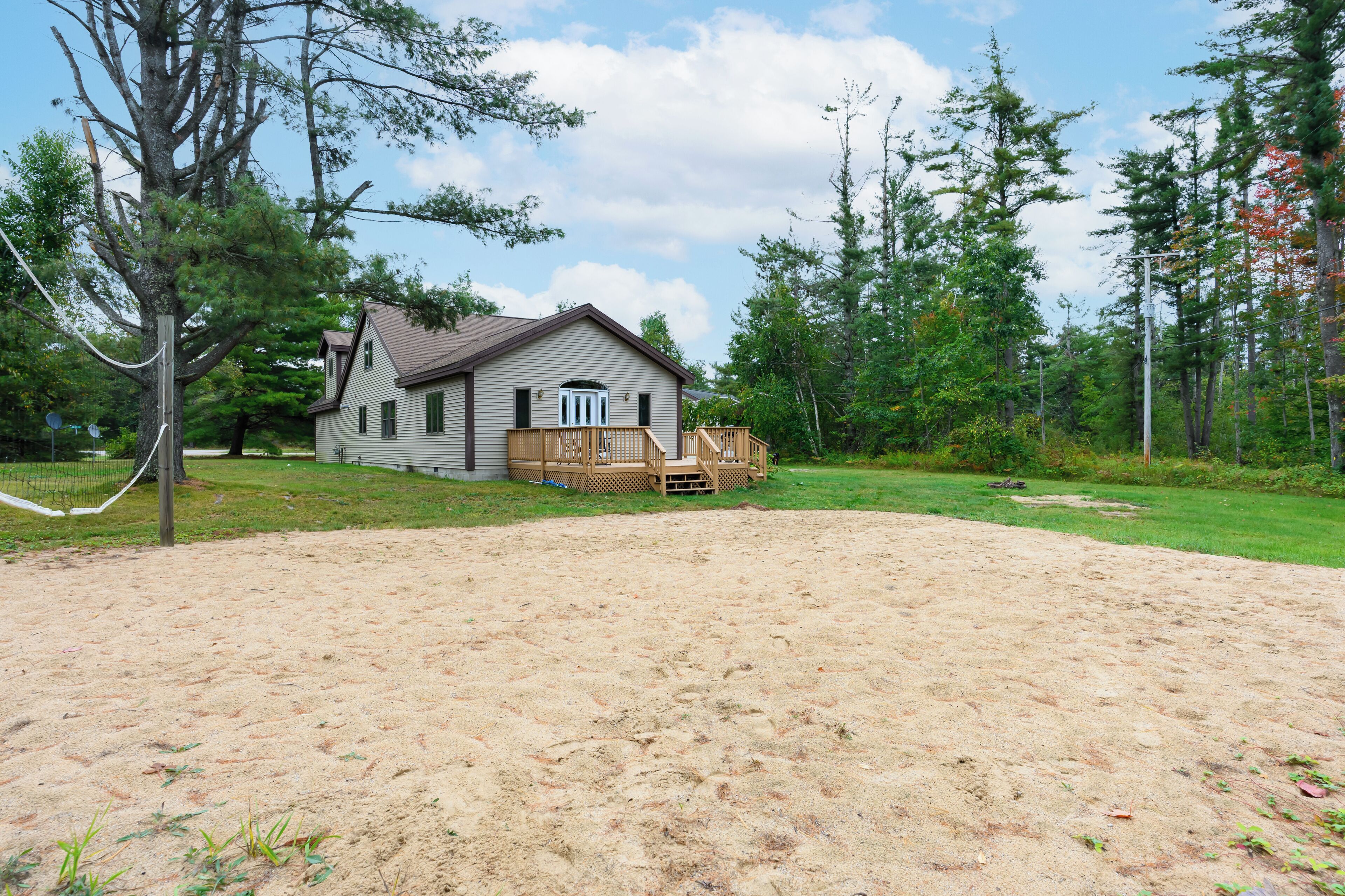 A house with a wooden deck, surrounded by a sandy area and trees.
