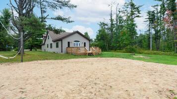 A house with a wooden deck, surrounded by a sandy area and trees.