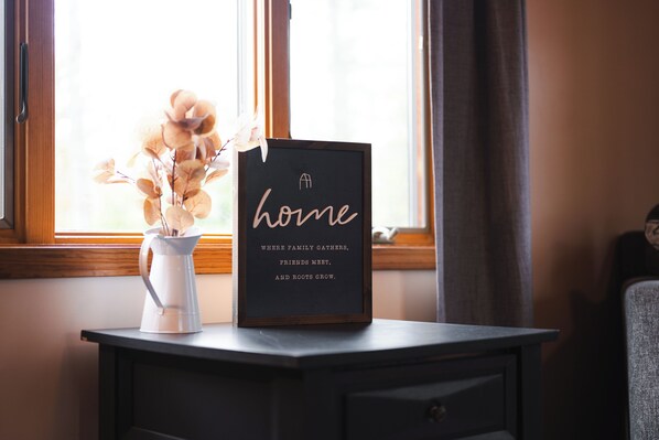 A framed sign with the word "home" and a quote on a dark surface near a window with a white vase containing flowers.