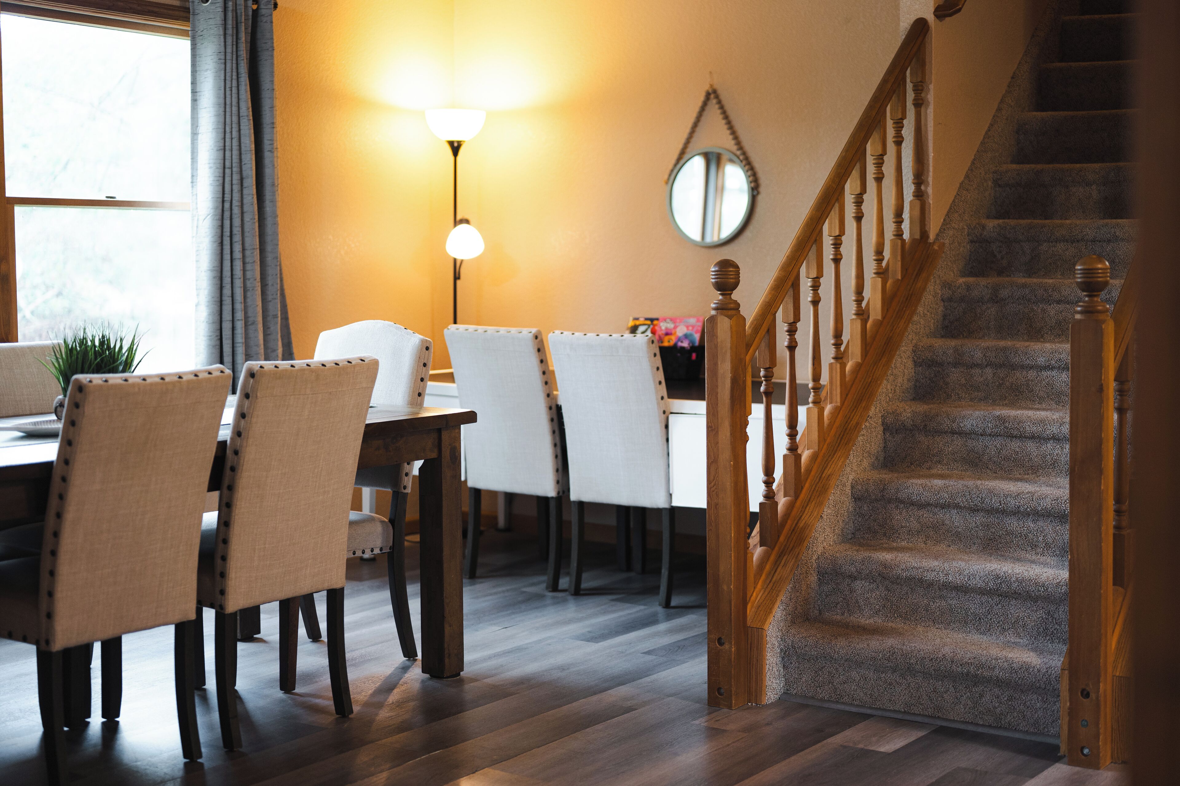 Un comedor con sillas y mesa de madera, una escalera con barandilla de madera y un espejo redondo en la pared.