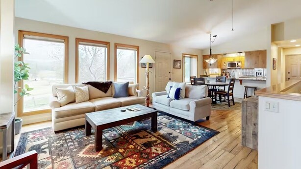 A spacious living area with two sofas, a coffee table, and a patterned rug. A ceiling fan and kitchen are visible in the background. - Peaceful Mountain View Home – Dog Friendly (Kalispell)