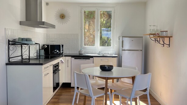 Een moderne keuken met witte kastjes, een centraal eiland, een eettafel met stoelen en een raam met uitzicht op groen.
