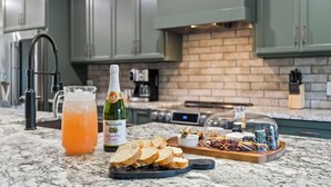 Un plan de travail de cuisine avec une carafe de jus d’orange, une bouteille de vin pétillant, une planche à fromages avec divers fromages et fruits, et une machine à café.