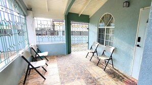 A covered outdoor area with tiled flooring, two white chairs, and a window with metal bars.