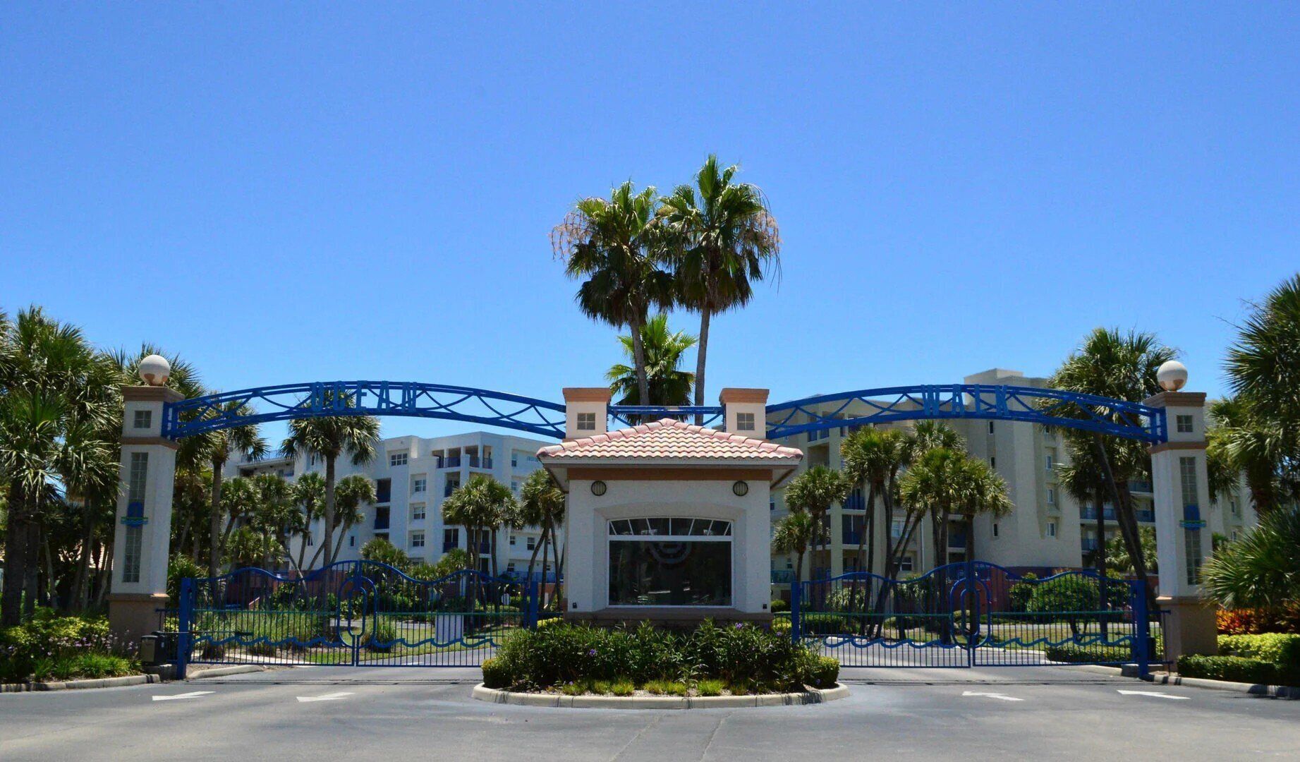 A gated entrance with a clear blue sky, palm trees, and a building with a red-tiled roof.