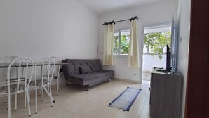 A small, well-lit room with a dining table and chairs, a gray sofa, a TV, and a window with curtains. - Pousada Village Maitinga (Bertioga)