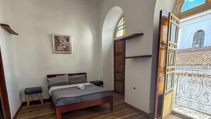 A bedroom with a wooden floor, a bed, a nightstand, a wall-mounted picture, and a door leading to an outdoor area with a view of a building. - Hotel Dubai (Cuenca)
