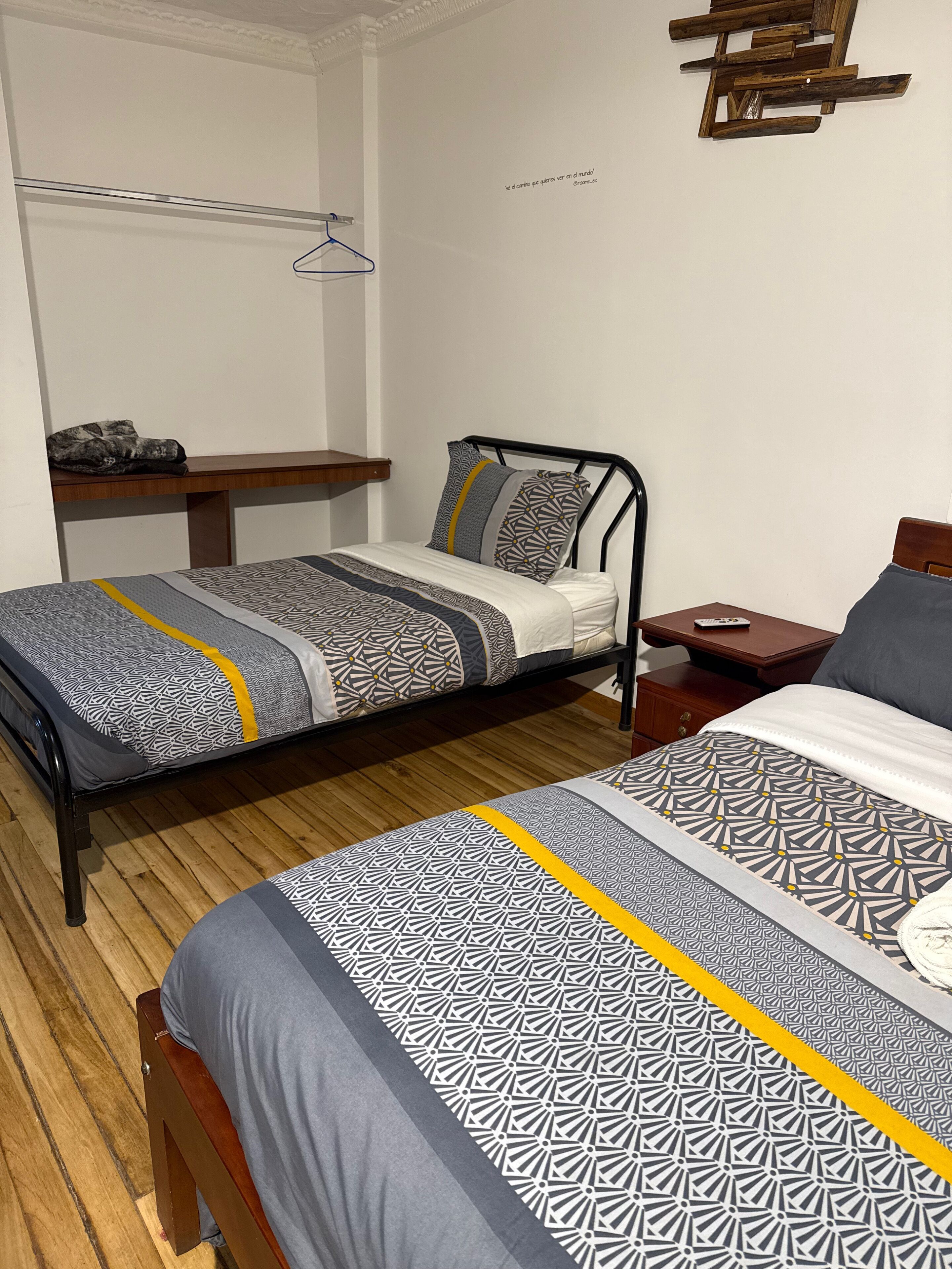 A bedroom with two beds, a wooden floor, a bedside table, a hanging light, and a wall-mounted shelf.