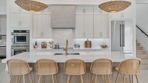 A modern kitchen with white cabinetry, a central island with bar stools, and two hanging wicker chandeliers. - NEW! Luxury 6BR Villa w/ Pool & Spa (Naples)