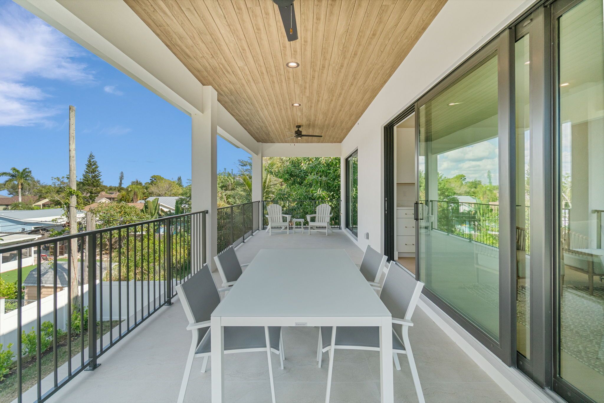 A modern outdoor dining area with a long table and chairs, a wooden ceiling, and a glass wall providing a view of the surrounding greenery and neighborhood.