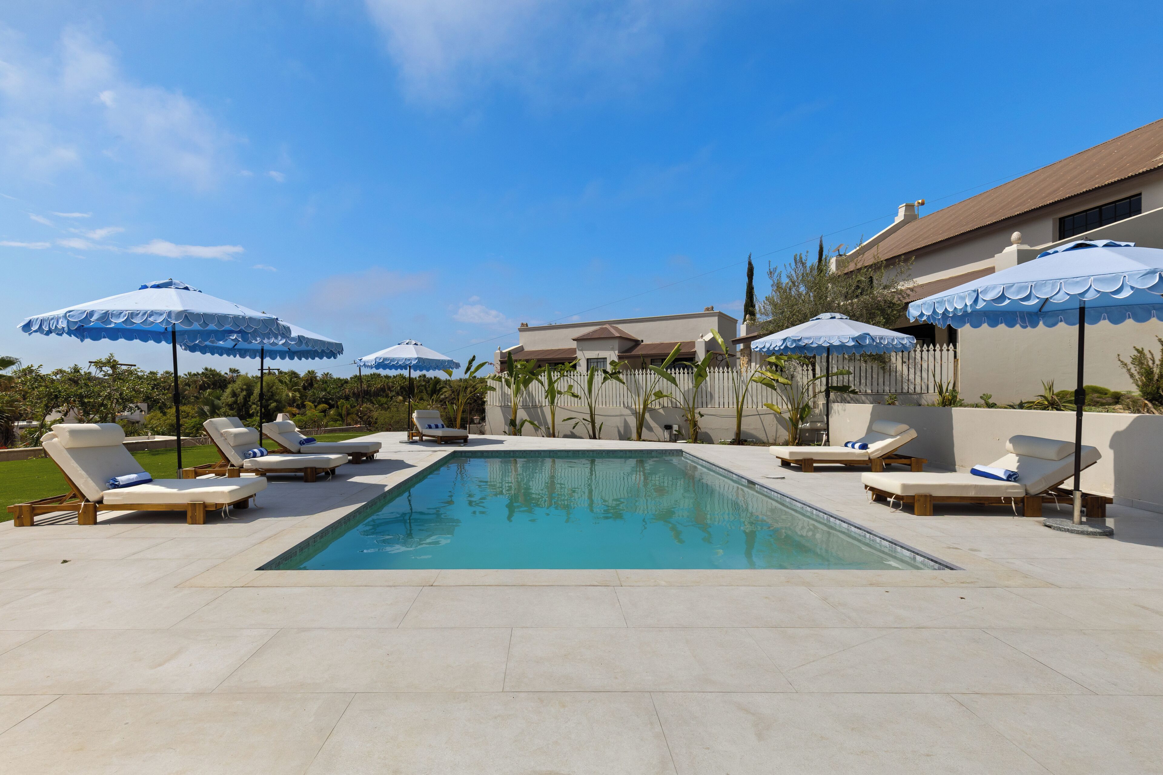 A swimming pool with lounge chairs and umbrellas, surrounded by a fence and greenery.