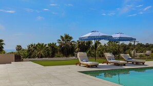 A pool area with lounge chairs and umbrellas, surrounded by palm trees and a clear sky. - Grand Villa del Mar (Todos Santos)