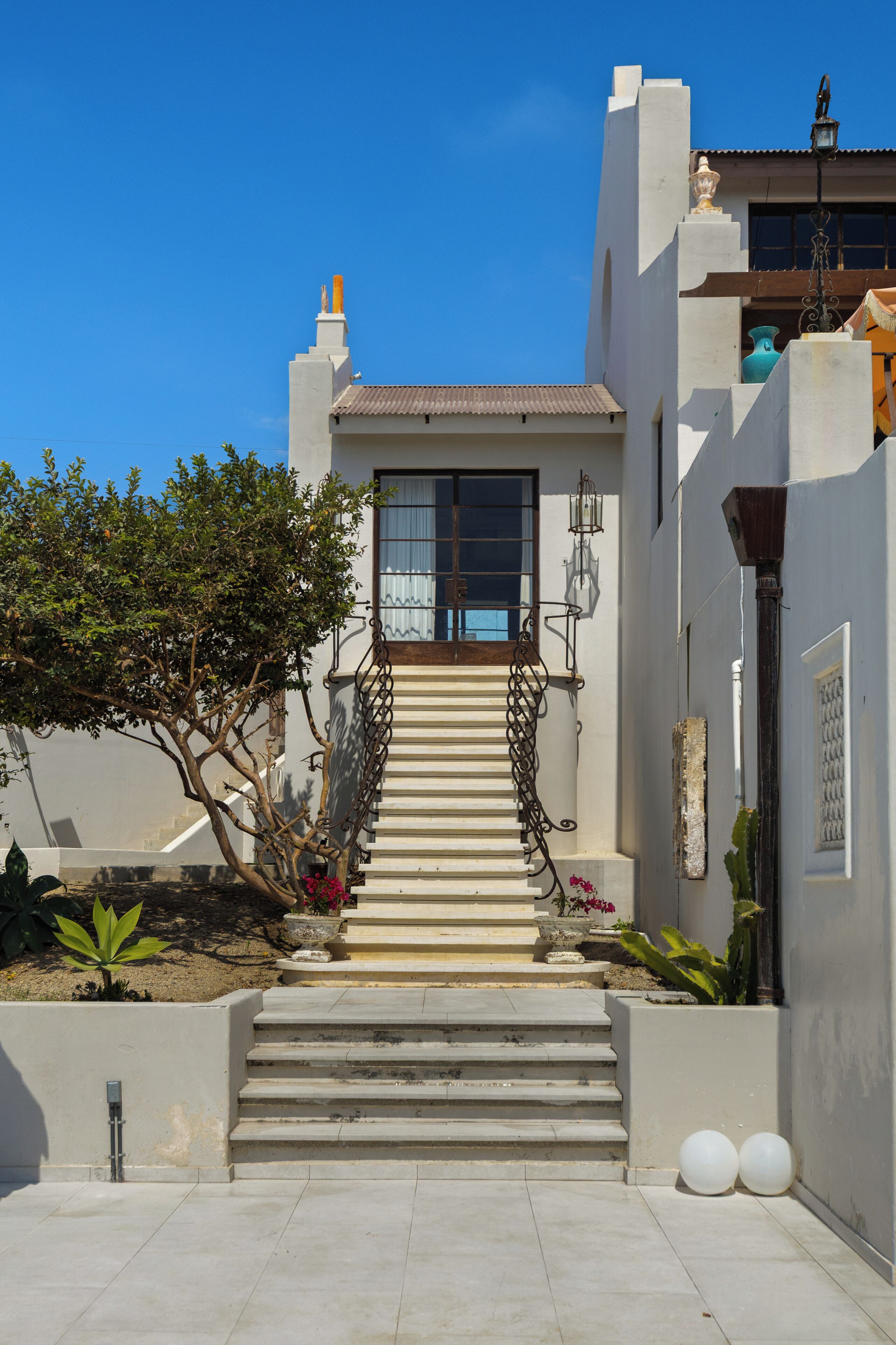 A building with a staircase leading to a door, surrounded by plants and a clear sky.