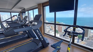 Un gimnasio con cintas de correr y una bicicleta estática, ventanas amplias con vista a la ciudad y al océano.