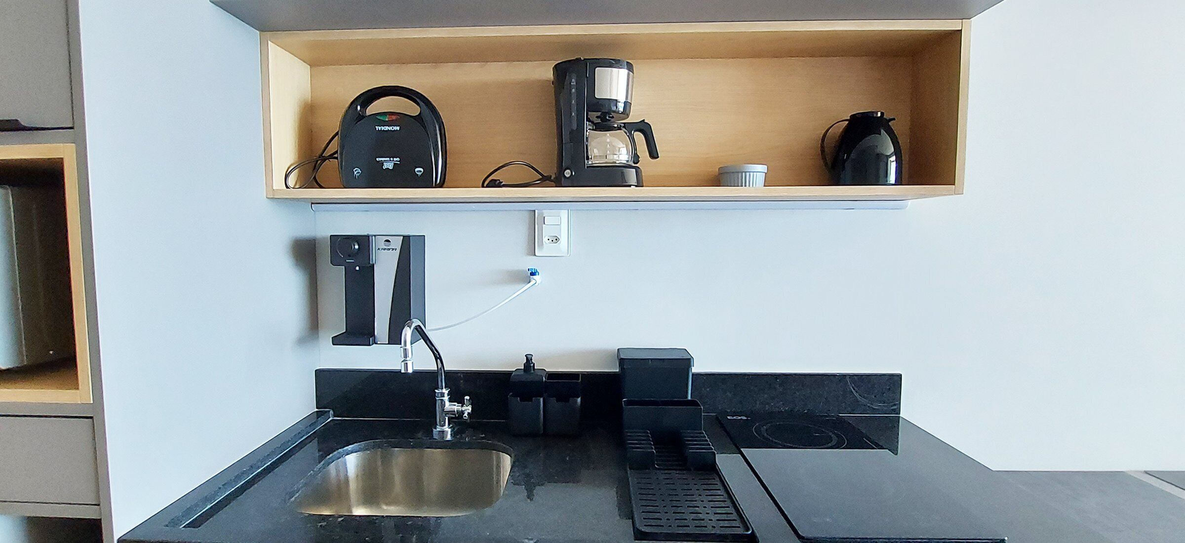 Una cocina con un lavamanos, una cafetera y un hervidor sobre una estantería.