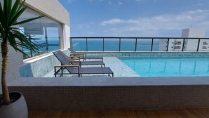 Una piscina en la azotea con sillones de descanso y una planta en maceta, con vistas a un paisaje urbano y al mar.