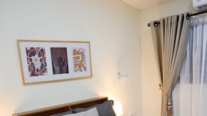 Een net opgemaakt bed met een grijze deken en kussens, een houten hoofdbord, een nachtkastje met een lamp, en een aan de muur bevestigde airconditioner.