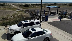 Un área de estacionamiento con coches blancos, un pasillo cubierto y vista a la playa.