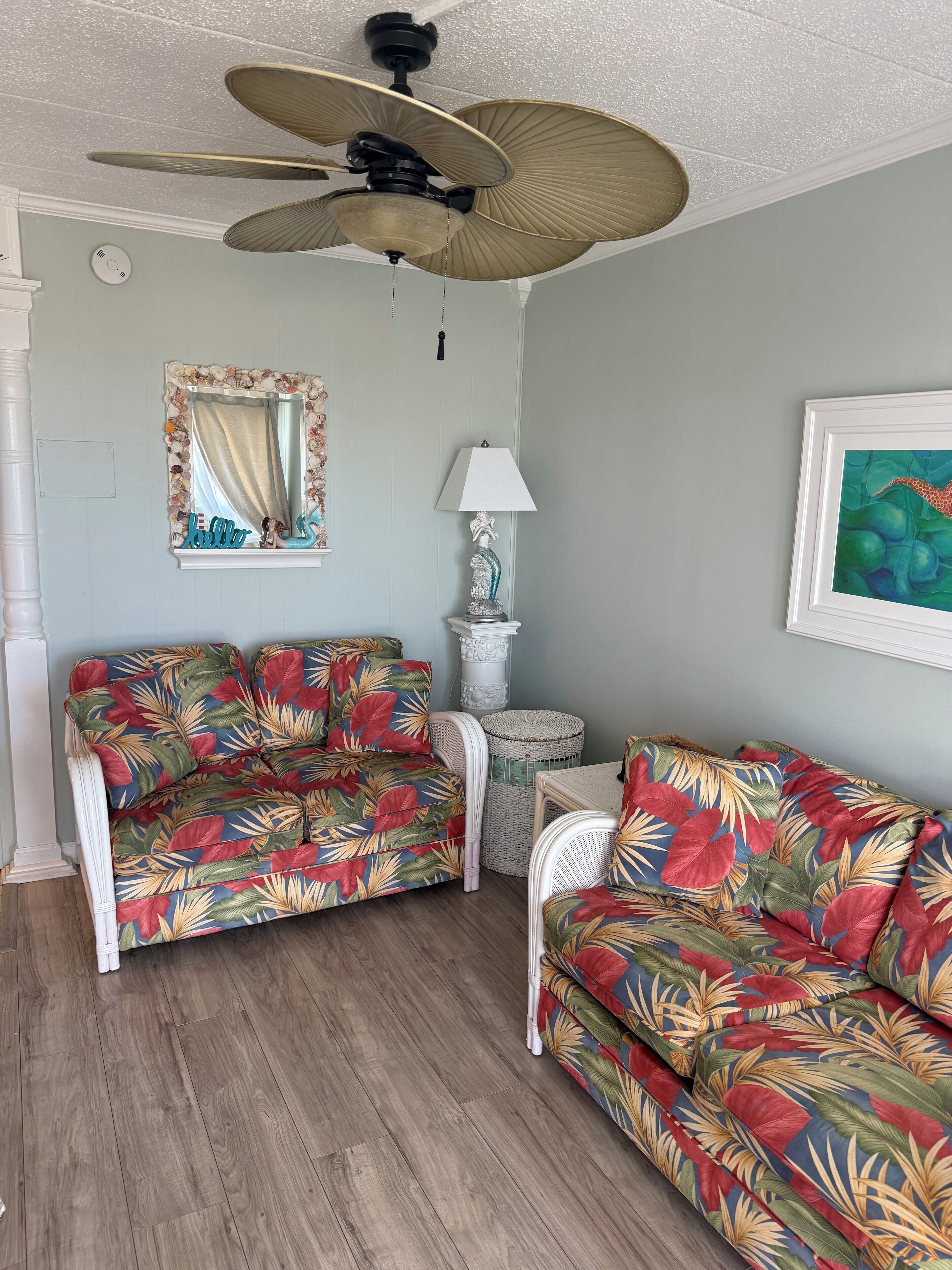 A room with two floral-patterned sofas, a ceiling fan, a mirror with a seashell frame, a lamp, and a framed painting.