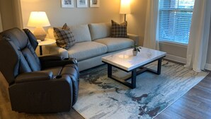 A living room with a sofa, a recliner, a coffee table, and a lamp. There are three framed pictures on the wall.