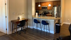 A modern kitchen with granite countertops, stainless steel appliances, and pendant lighting. There are three bar stools and a dining table with chairs.
